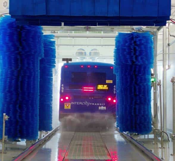 The back of a blue bus going through an automatic truck wash system with blue brushes.
